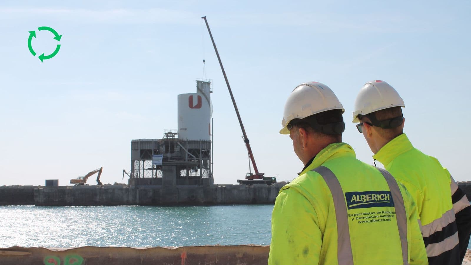 Two Alberich technicians watch a silo being demolished on a quayside, with a mobile crane and an excavator