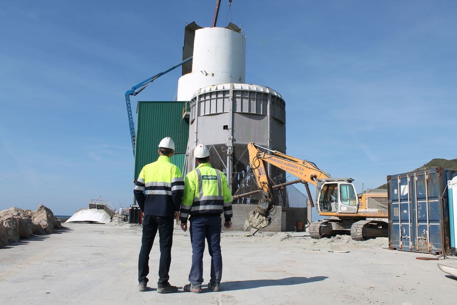 Two technicians in helmets and high-visibility vests on an industrial dismantling site, with an excavator in the background
